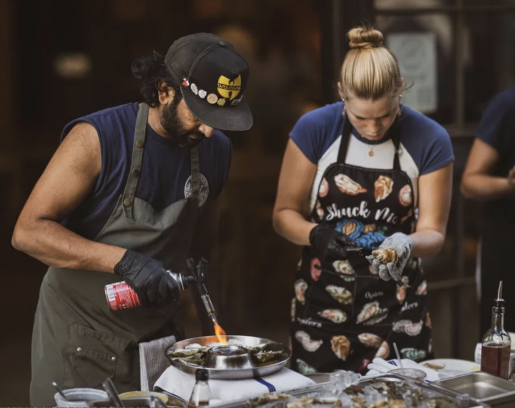Two shuckers preparing fresh oysters over an open flame at an outdoor station